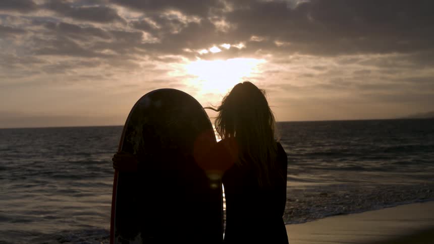 A silhouetted surfer stands on the beach overlooking the ocean at sunset