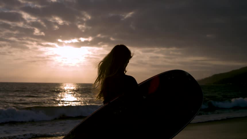 Woman carries her surfboard across the beach as the sun shines through clouds on the horizon