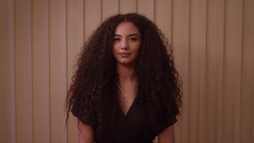 A young woman with curly hair is featured in a calm centered medium closeup shot indoors maintaining a neutral expression and direct eye contact with the viewer against a simple light brown background composed of vertical slats