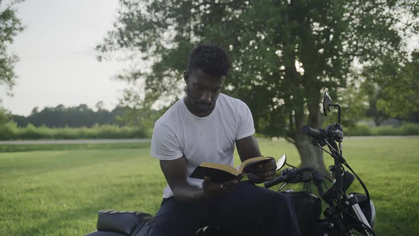 Man sits on his motorcycle in a field reading a book