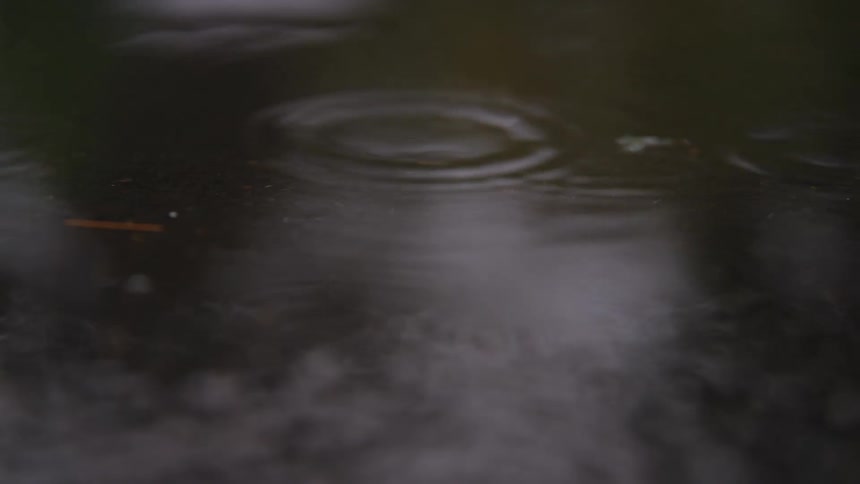 A closeup shot captures raindrops hitting a puddle on the ground creating expanding ripples on the dark wet surface