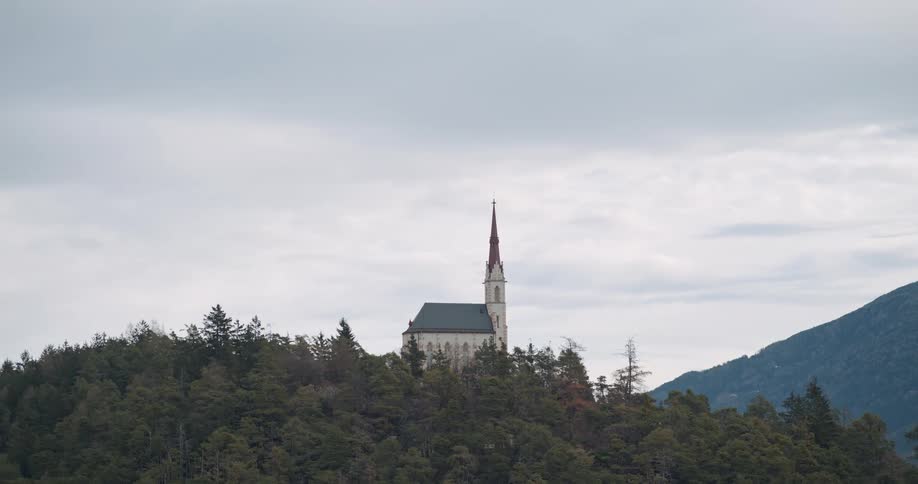 Aerial shot of a church with a small tower in the middle of a forest on top of a large hill