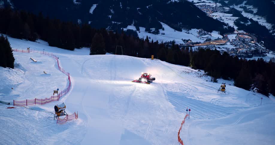 Snow groomer lights up the slope at dusk in the snowy mountains