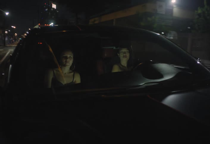 Two women drive through a city at night as seen through the front windshield of their car