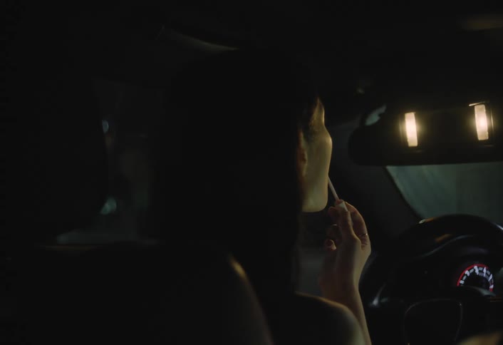 Young woman sitting in the car at night looks at herself in the visor mirror and applies makeup