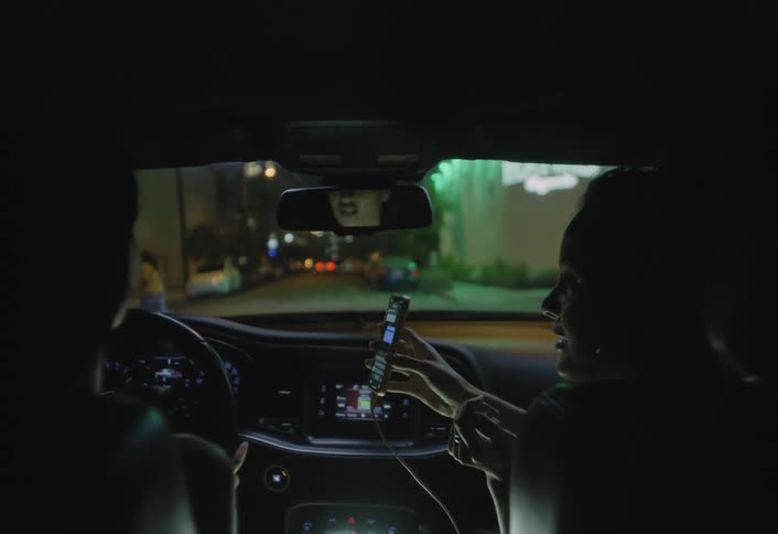 Female passenger shows her smartphone to the man driving while they pass through the city at night