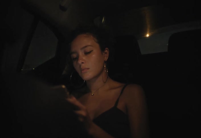 A young woman with her hair pulled up wearing large hoop earrings and a black top is seated in the back of a dimly lit car at night where she is seen holding and looking intently at a smartphone in her hands as the vehicle drives along a street