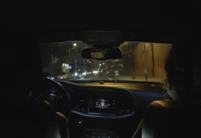 Two people inside a car are talking while driving down a city street at night