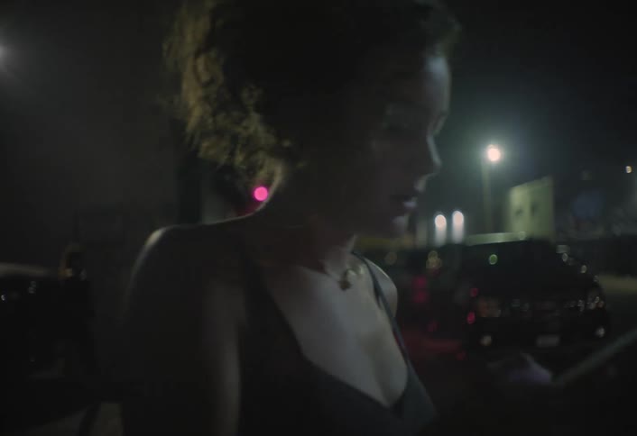 The video set at night in an alley or parking area begins with a long shot of people walking away from a buildings brightly lit red doorway towards parked cars it then cuts to a closeup of a woman in a dark dress checking her phone and taking a puff