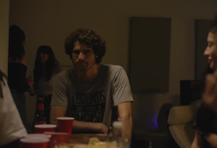 A woman with large hoop earrings reaches toward a table with a red cup chips and a water bottle as friends sit nearby in dim light