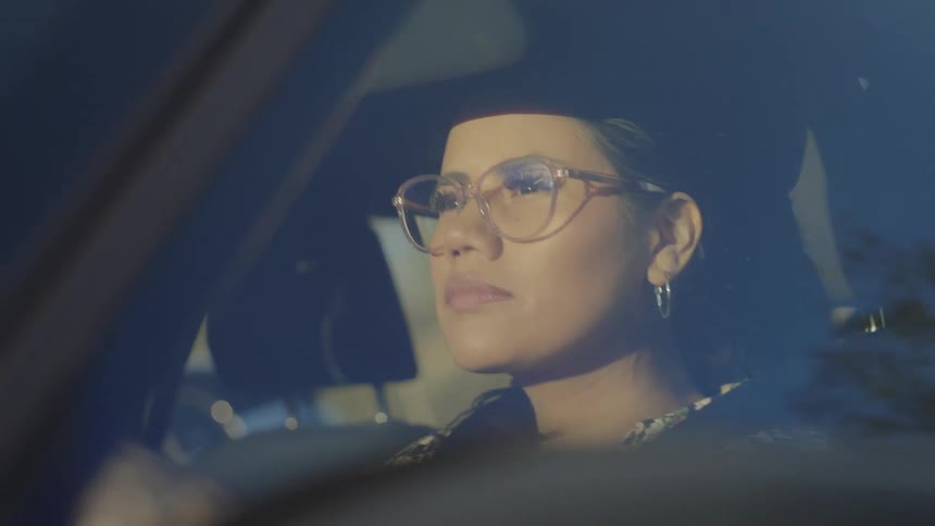 The video is a closeup shot of a woman with glasses sitting in the drivers seat of a car she stares straight ahead with a neutral perhaps contemplative expression lit softly by the interior and external light