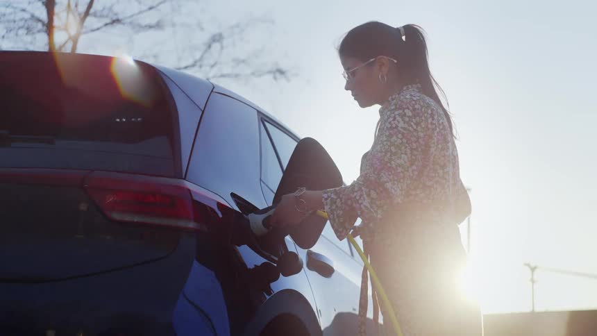 Young woman wearing glasses pumping fuel into a vehicle outdoors during dusk or dawn