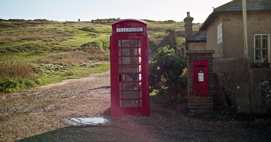 old Red telephone booth next to a building at the foot of a hill covered with grass and plants
