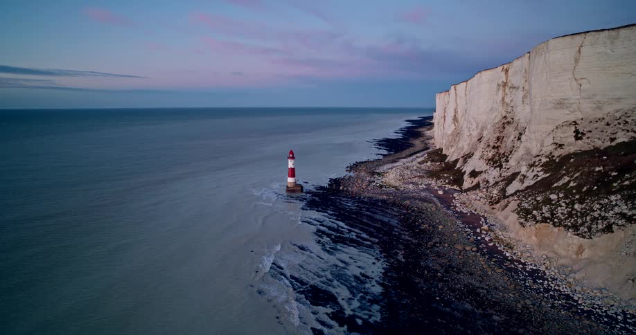 A red and white lighthouse is by the cliff on the shore at sunset