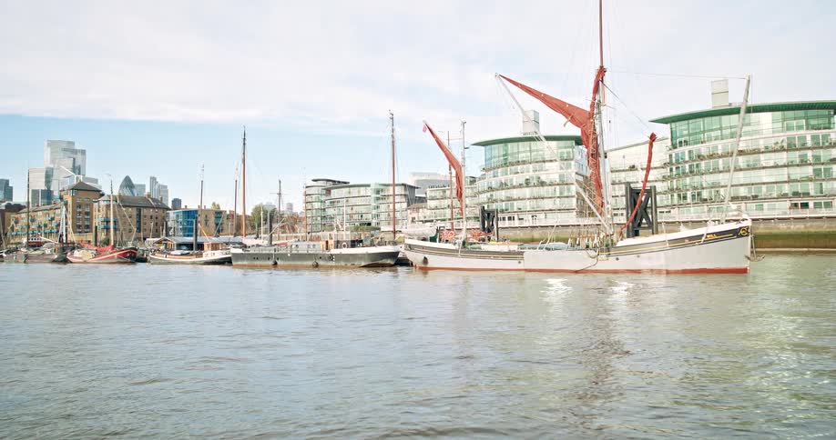 Multiple ships are docked along the thames river in england, office buildings behind