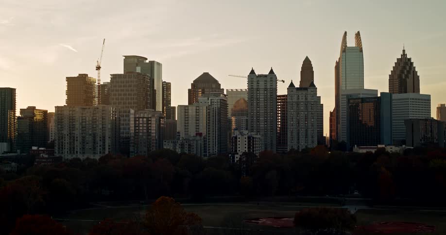 The video is a timelapse of a city skyline at sunset featuring a collection of highrise buildings and skyscrapers silhouetted against a bright hazy sky in the dark foreground there is an area of dense trees and a park that runs along the bottom of t