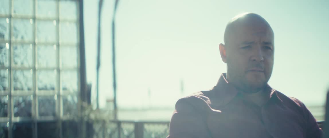 This video features a man the same person from the previous clip bald wearing a reddishbrown shirt sitting outdoors at a small table while looking at a laptop he is in a very bright sunlit location possibly a balcony or patio indicated by a deco