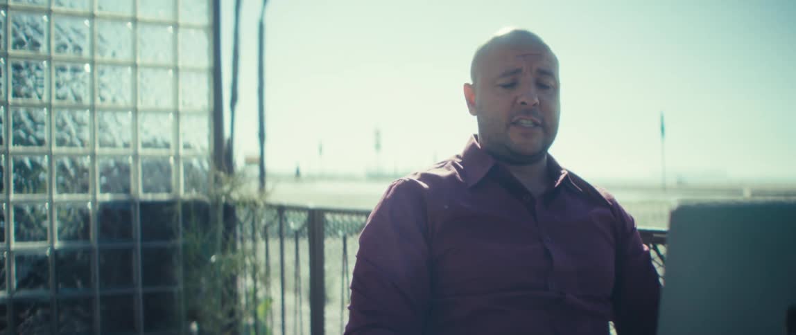 A man speaking on a call while using a laptop computer at a table outdoors during daytime