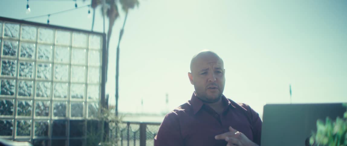 A man sitting at a table on a ground floor balcony speaks and gestures in front of an opened laptop during a video call