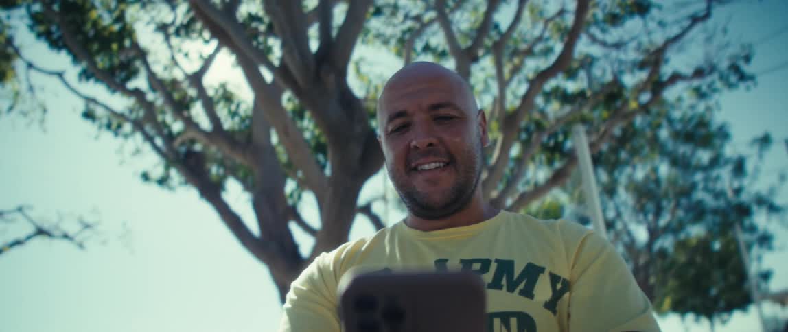A young man wearing a tshirt that says army veteran records a selfie video in the park under a tree at midday