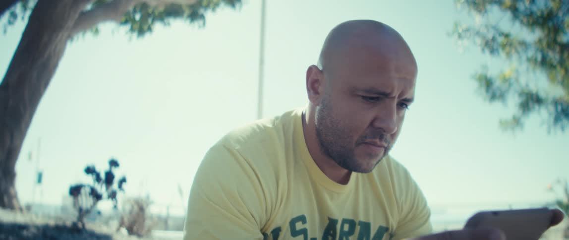 Young bald man wearing a yellow shirt frowning while looking at a smarphone on his hands outdoors during day time