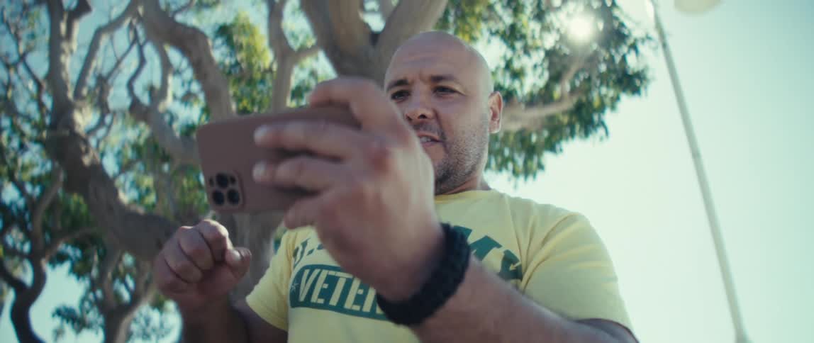 A man standing under a tree looks serious as he watches a video on his phone then suddenly laughs and pumps up his fist in celebration