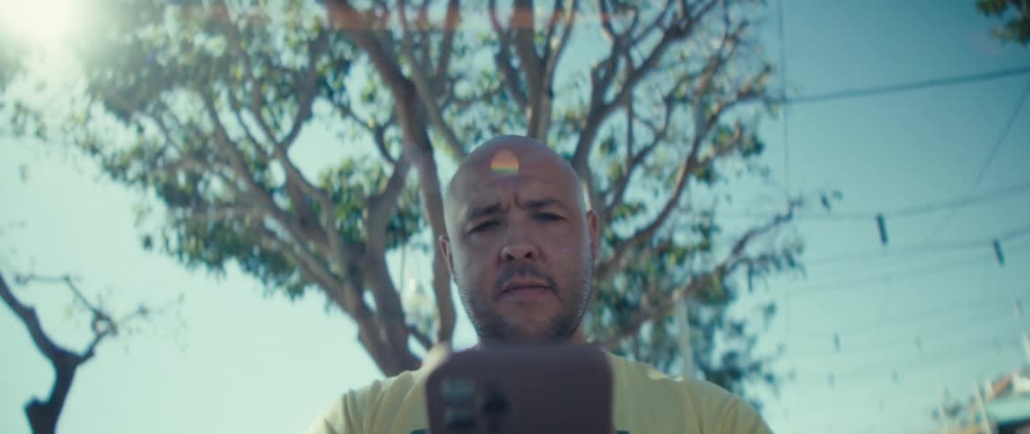 A young man wearing a yellow tshirt walks down the street using his mobile device with trees lining the street on a sunny day