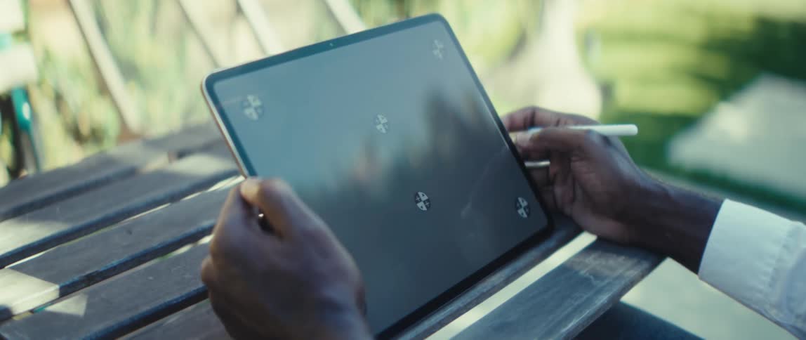 A closeup shot focuses on a persons hands holding a large tablet with a blank dark screen that features onscreen tracking markers the person is seated outdoors on a wooden bench or table and is holding a stylus near the screen appearing ready to dra