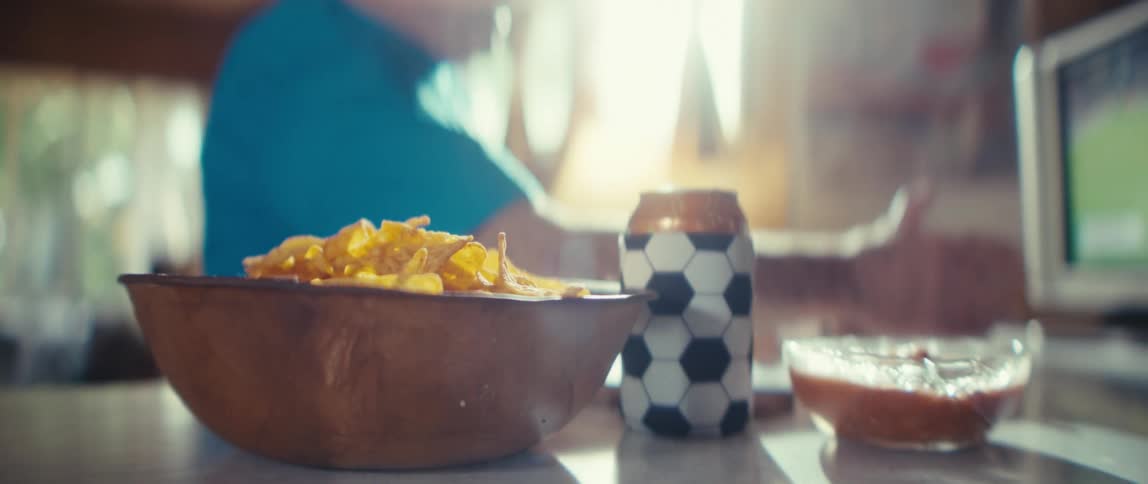 A bowl of chips and salsa with a soccerpatterned drink can sit on a countertop as a elderly man watches a screen in warm sunlight