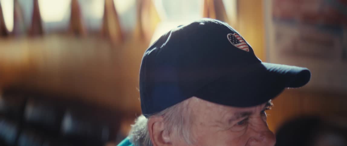 Elderly man in a blue shirt and dark cap smiles