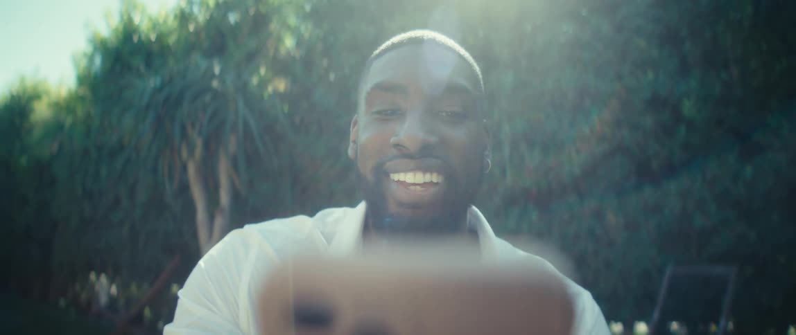 Bearded black man wearing a white shirt talking and smiling at a smartphone on his hands outdoors during day time