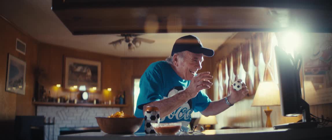 Elderly man wearing a soccer shirt and holding a small football on his hand cheering in front of a screen indoors during day time