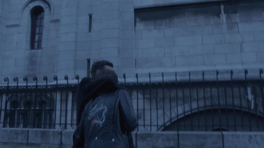 A young couple with their arms around each other talks and laughs as they walk down a street in paris near the metal fence in front of a stone building