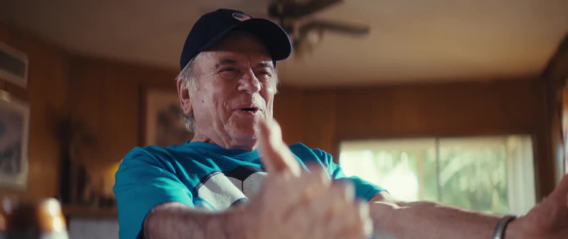 An elderly man wearing a soccer fan t shirt a cap with an american flag and holding a small soccer ball celebrates while watching a game on television