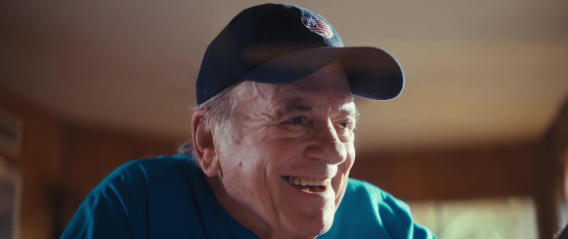 An elderly man wearing a soccer fan t shirt laughs and claps excitedly as he watches television