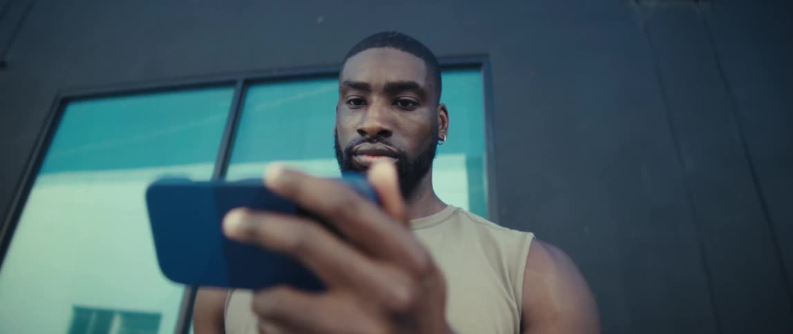 A man with a full beard and a high fade haircut wearing a sleeveless beige shirt stands outside a dark building with large reflective windows looking down at a mobile phone he holds horizontally he slowly raises his gaze from the phone to look intentl