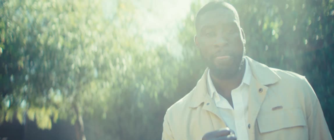 A stylish man dressed in a light beige jacket and dark trousers is shown in a closeup while playing miniature golf outdoors on a bright day initially focusing on his putt before looking up at the camera with a celebratory smile