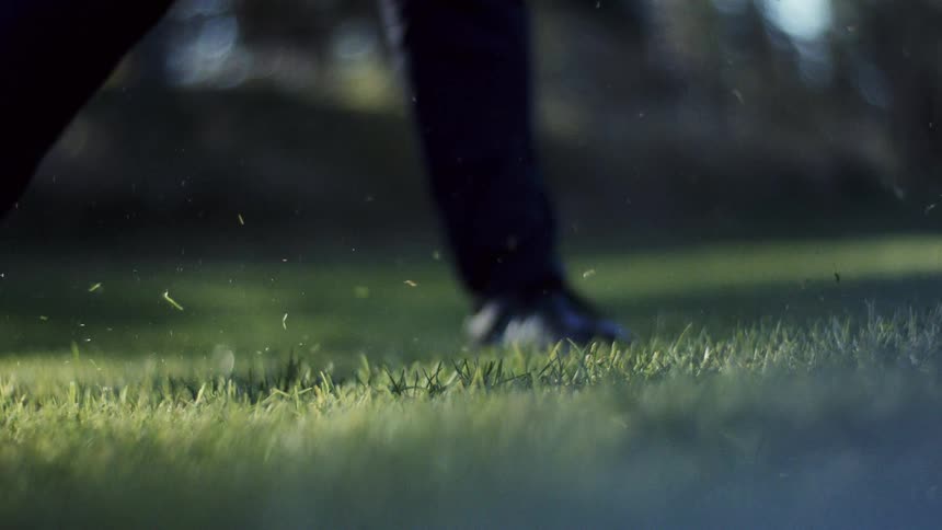 A lowangle slowmotion shot captures a golf club hitting a ball off the grass sending a large divot flying into the air