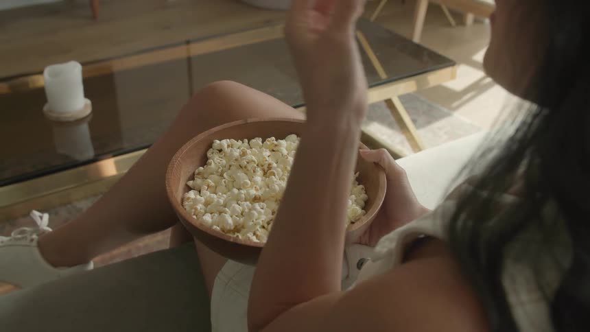A woman sitting in living room eating from a large bowl of popcorn on her lap