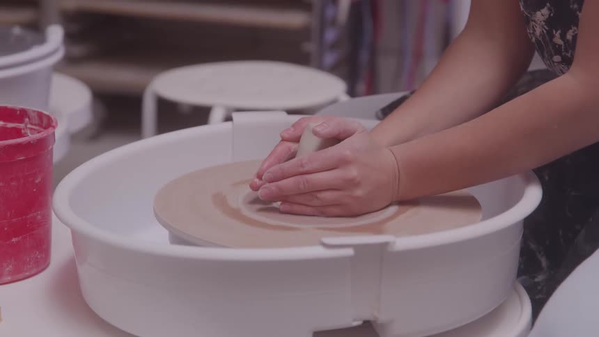 A woman focuses intently as she shapes clay on a pottery wheel in a studio surrounded by tools and pottery equipment