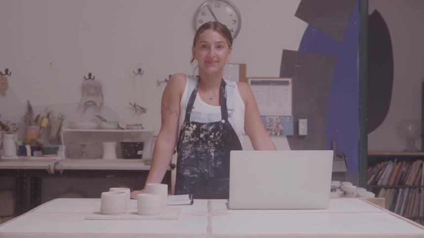 A woman wearing an apron stands next to some unfinished ceramic cups