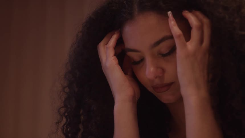 A closeup shot features a young woman with long curly dark hair and expressive eyes she brings her hands up to her head and gently bows her head down conveying a feeling of distress exhaustion or deep thought