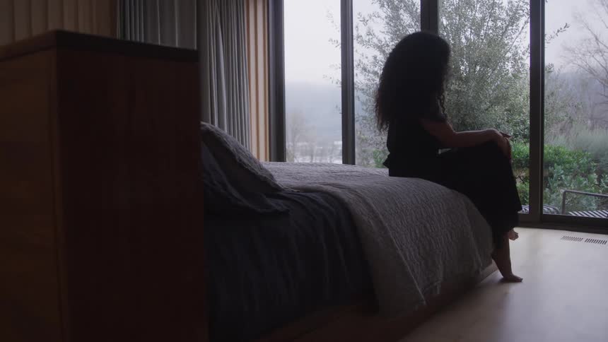 A young black woman sits at the edge of the bed in a bedroom looking out the window as the large glass panes reveal green vegetation outdoors at midday