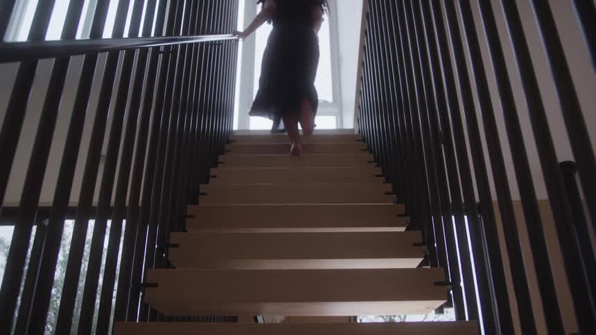 Woman with long dark hair wearing a long dress walks up the stairs inside a building with a wall of windows