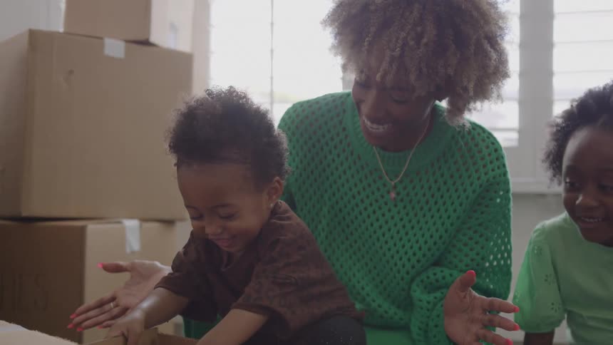 Mother and her child play with a small box while her daughter smiles and looks on near a stack of packed boxes