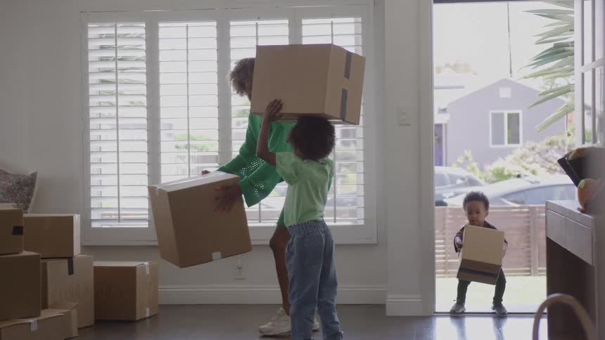 An adult in a bright green outfit sets down a heavy cardboard box assisted by two young children who are playfully carrying smaller boxes into a bright whitewalled room