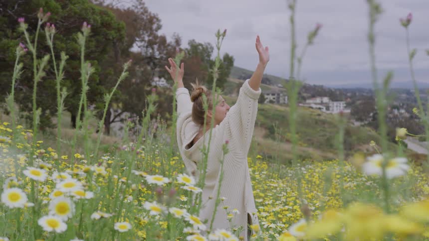 A woman wearing a white dress is raising her arms upwards and she is in a field with yellow and white flowers