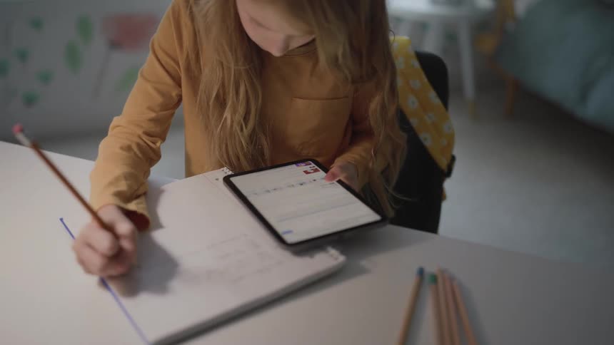 Girl writing in a notebook while looking at a tablet