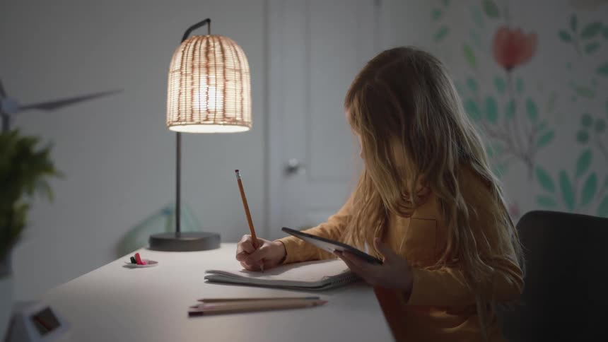 A young girl writes with a pencil while also holding a tablet computer