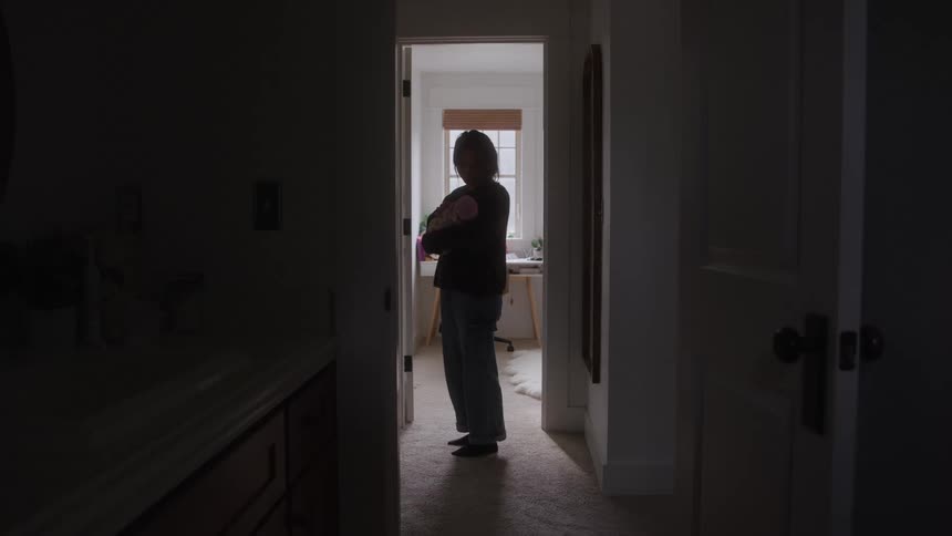 A silhouette of a person standing in a doorway is framed by the darkness of the foreground hall and the light from the room behind them the person appears to be holding an infant suggesting a moment of quiet care or transition